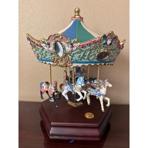 May include: A decorative carousel music box featuring a dark wood hexagonal base, a canopy with blue and green panels, and gold filigree accents. Three painted horses in black, white, and cream with floral details are suspended from the canopy.