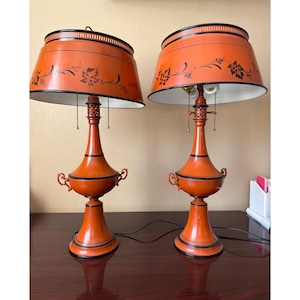 May include: Pair of orange table lamps with black trim and floral designs on the shades. The lamps have a classic, elegant shape with decorative handles and a detailed base. The shades are a warm orange color, and the lamps are set on a dark wooden surface.