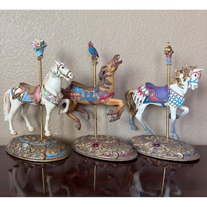 May include: Three carousel horse figurines mounted on gold-colored poles and ornate oval bases. The left horse is white with pink floral accents, the center is brown with a blue parrot topper, and the right is white with blue and red saddle details.