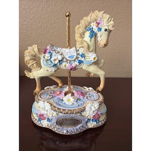 May include: A cream-colored carousel horse figurine with a gold-tone central pole, adorned with blue, pink, and white floral accents and pearl-like beads. The base features the text 20th Anniversary San Francisco Music Box Company.