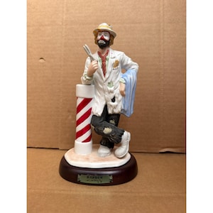 May include: A ceramic figurine of a barber clown, wearing a white lab coat and holding scissors. The clown leans against a barber pole with red and white stripes. The base of the figurine has the word "BARBER" on it.