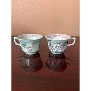May include: Two pale green porcelain teacups with handles. The left cup features a single floral branch. The right cup displays a detailed scene of a bird perched on a flowering tree. Both cups have a geometric red and white pattern along the rim.