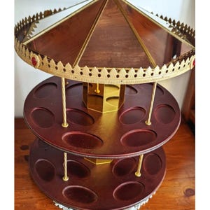 May include: A two-tiered carousel-style display stand featuring dark wood circular shelves with oval indentations. The structure is supported by gold-toned metal pillars and topped with a decorative gold-toned metal canopy rimmed with small red accents.