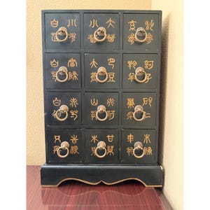 May include: A black wooden apothecary chest featuring twelve individual drawers arranged in a four-by-three grid. Each drawer is adorned with gold-colored Asian characters and a circular brass pull handle. The base has a decorative cutout design.