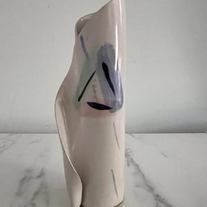Puede incluir: Un jarrón de cerámica blanco roto con un diseño único y plegado. El jarrón presenta detalles abstractos pintados en azul, negro y rosa. El jarrón mide aproximadamente 25 cm de alto y tiene una base redondeada.