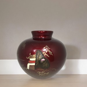 Op de afbeelding: Een rode en gouden keramische vaas met een rond lichaam en een geschilderd ontwerp van een berg en een gebouw. De vaas staat op een lichtbruin houten oppervlak.