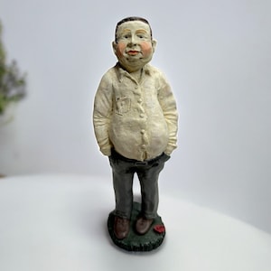 May include: A figurine of a person with a white shirt and gray pants standing on a green base. The person has a round face and a friendly expression.