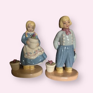 May include: Two ceramic figurines of children in Dutch attire. The girl wears a blue dress and holds a flower pot. The boy has a light blue jacket and pants, and holds a hat. Each figure stands on a round base.