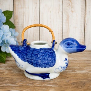 May include: A ceramic teapot shaped like a duck, featuring a white body with blue painted feather details and a blue head. The teapot includes a curved, light brown woven rattan handle attached to two metal loops on the top of the duck's back.