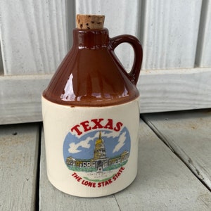May include: A brown ceramic jug with a cork stopper and a white label that says "Texas The Lone Star State" with a drawing of the Texas State Capitol building in Austin.