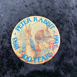 Puede incluir: Un botón redondo de color dorado con una imagen de dos conejos en un jardín. El botón dice "Peter Rabbit 1893 - 1993 100 Years" en letras azules.