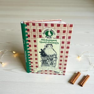 May include: A cookbook titled "Old-Fashioned, Country Christmas" by Gooseberry Patch. The cover features a Santa Claus illustration on a red and white plaid background. The book is spiral-bound with a green spine. Two cinnamon sticks and a string of lights are next to the book.