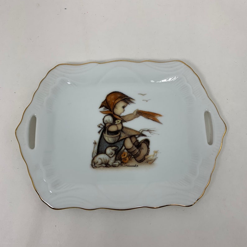 Op de afbeelding: Een wit porseleinen dienblad met een gouden rand heeft een meisje met een mand en twee lammetjes. Het meisje draagt een bruine jurk en een rood-witte muts. Het dienblad heeft twee handvatten aan de zijkanten.