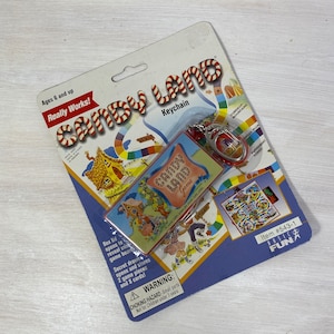 Può includere: Un portachiavi Candy Land con un design a forma di bastoncino di zucchero a strisce rosse e bianche. Il portachiavi include una piccola plancia di gioco che si apre per rivelare una plancia di gioco in miniatura, pedine e carte. La confezione include il testo "Really Works!" e "Item #543-1".