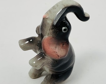 Può includere: Una piccola statuetta di elefante in ceramica con un corpo grigio e nero, orecchie rosa e un occhio nero. L'elefante è in piedi sulle zampe posteriori con la proboscide alzata.