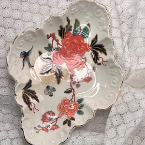 May include: A white ceramic dish with a scalloped edge and gold trim, featuring a central floral design with coral-colored peonies, dark blue leaves, a small blue bird, and a butterfly, all set against a smooth, glossy, off-white surface.