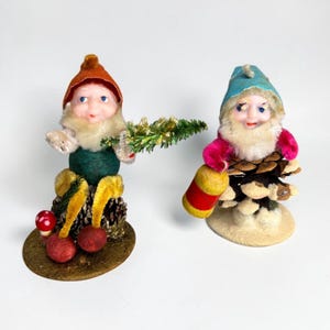 May include: Two gnome figurines with white beards. The left gnome wears an orange hat, green shirt, and yellow pants, holding a green pine branch. The right gnome wears a blue hat and pink sleeves, holding a yellow and red barrel on a pinecone base.