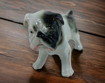 Può includere: Statuetta di bulldog in ceramica in posizione eretta. Il cane ha un corpo bianco con muso e orecchie neri e grigi. La bocca è aperta, rivelando una lingua rosa. La statuetta è su una superficie di legno.