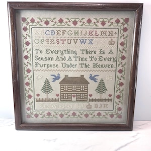 May include: A framed cross-stitch sampler featuring a dark wood frame, a floral border, the alphabet, and the text: To Everything There Is A Season And A Time To Every Purpose Under The Heaven. The design includes a house, trees, and birds.