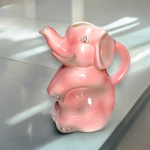 Op de afbeelding: Een roze keramische olifantenkan met een verhoogde tuit en een handvat. De kan heeft een glanzende afwerking en een speelse uitdrukking.