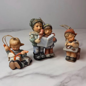Pode incluir: Três figuras de cerâmica com laços dourados para pendurar. Uma criança está sentada com um presente rotulado 'To a good girl', duas crianças de pé seguram um livro intitulado 'Heavenly Trio', e uma criança trabalha numa pequena secretária.