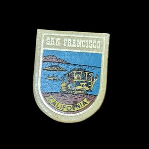 Puede incluir: Pin de recuerdo rectangular con una escena del tranvía de San Francisco. El pin tiene un borde dorado con las palabras "SAN FRANCISCO" y "CALIFORNIA". La imagen muestra un tranvía amarillo, agua azul y tierra marrón.