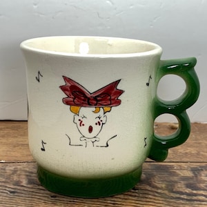 May include: A white ceramic mug with a green handle and a green rim. The mug features a hand-painted design of a person with a red bow singing with musical notes.