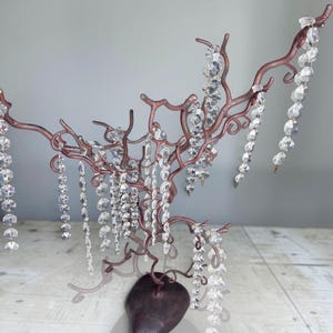 Puede incluir: Un soporte para joyas de metal color cobre con forma de árbol y una base oscura redondeada. Las ramas están adornadas con múltiples hilos colgantes de cuentas de cristal transparentes y facetadas que cuelgan de las ramas metálicas.