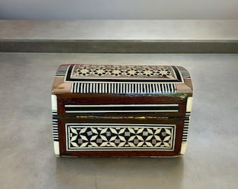 May include: A small wooden box with intricate inlaid geometric patterns in shades of brown, white, and black. The box has a hinged lid and is decorated with a variety of geometric shapes.