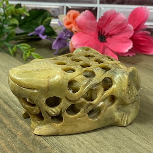 Puede incluir: Una figurita de rana de piedra tallada a mano con un diseño detallado e intrincado. La rana está hecha de una piedra marrón claro con un tono ligeramente verde. La figurita es hueca y tiene un patrón de celosía en toda su superficie.