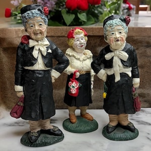 May include: Three ceramic figurines of women wearing black dresses and white collars. The women have white hair and are holding flowers. The figurines are standing on green bases.