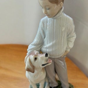 May include: A porcelain figurine of a boy in a white sweater and brown pants standing with a white dog. The boy is looking down at the dog.