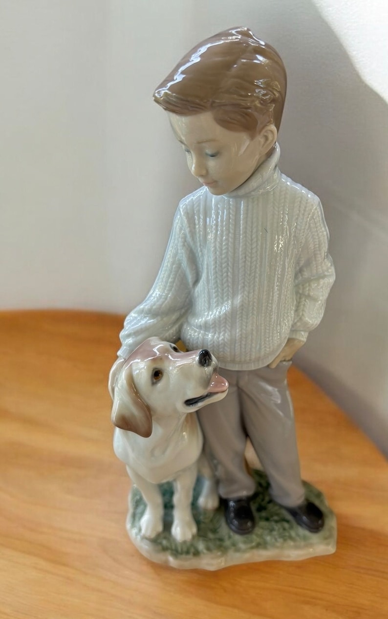 May include: A porcelain figurine of a boy in a white sweater and brown pants standing with a white dog. The boy is looking down at the dog.