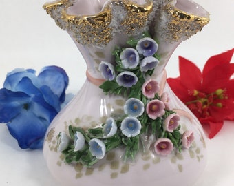 May include: A small, pink porcelain vase with a ruffled top and gold trim. The vase is decorated with 3D flowers in shades of pink, blue, and white.