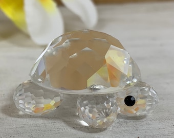 May include: A clear crystal turtle figurine with a faceted shell and legs. The turtle's shell is a light tan color, and it has a black eye. The figurine is a decorative item.