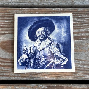May include: Square ceramic tile featuring a blue and white portrait of a man in a hat and ruffled collar. The artwork is reminiscent of Delftware pottery, with intricate details and a vintage aesthetic.