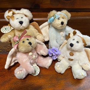 Pode incluir: Quatro ursos de pelúcia com asas de anjo. Os ursos estão vestindo roupas de diferentes cores, incluindo rosa, azul e branco. Os ursos têm faixas de cabeça douradas e brancas com flores. Os ursos estão sentados em uma superfície de madeira.
