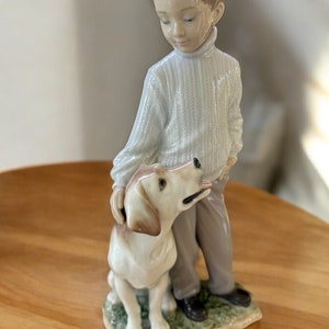 May include: A porcelain figurine of a boy with brown hair wearing a white sweater and brown pants standing next to a white and brown dog. The figurine is set on a green base.