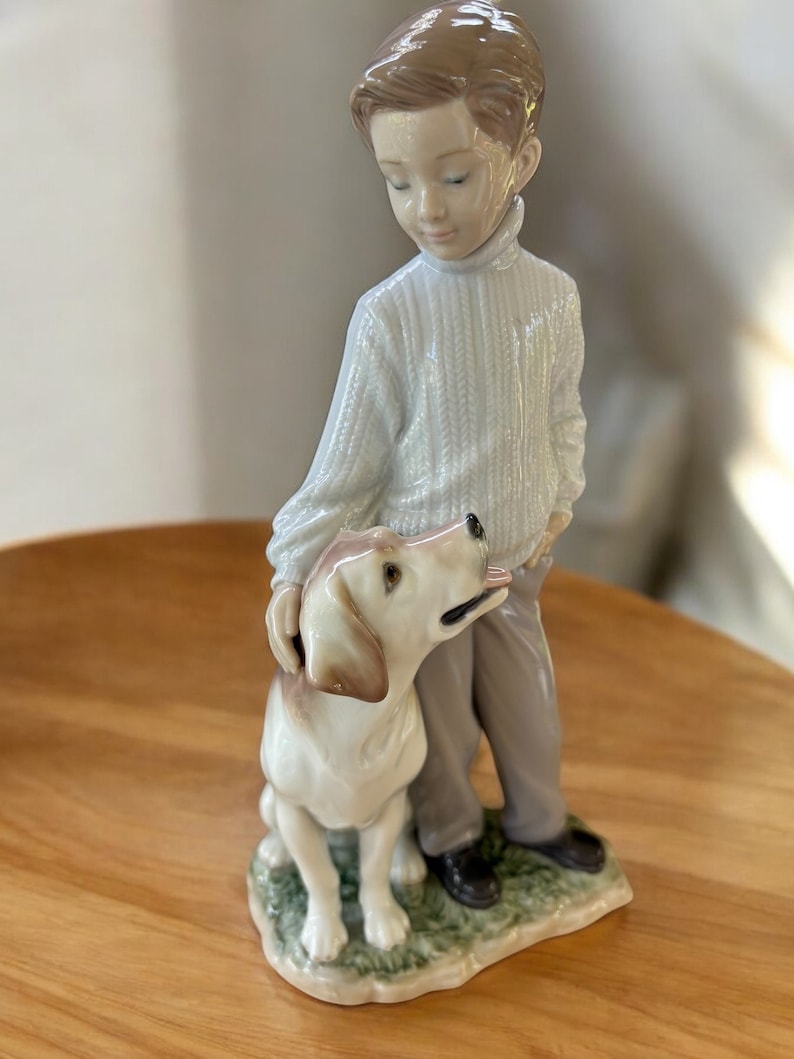 May include: A porcelain figurine of a boy with brown hair wearing a white sweater and brown pants standing next to a white and brown dog. The figurine is set on a green base.