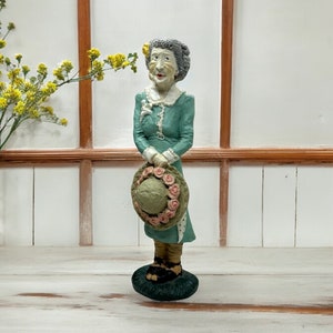 May include: A figurine of a woman with gray hair wearing a green dress and holding a straw hat with pink flowers. The figurine is standing on a green base.