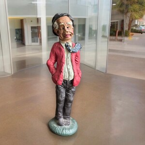 May include: A figurine of a man wearing a red jacket, a white shirt, black pants, and a blue and white patterned tie. The figurine is standing on a green base.