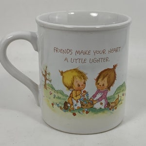 Könnte beinhalten: Weiße Keramiktasse mit einer bunten Illustration von zwei Kindern, die Blumen pflücken, und dem Text "Friends make your heart a little lighter."