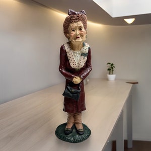 May include: A figurine of an older woman with red hair, wearing a burgundy dress with white polka dots, a white blouse with a white flower, and black shoes. She is holding a black purse and has a friendly expression.