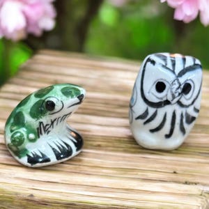 May include: Two ceramic ornaments: a green frog with black detailing and a white owl with black markings. The frog has green spots and the owl has a detailed face. Both are on a wooden surface, with pink flowers in the background.
