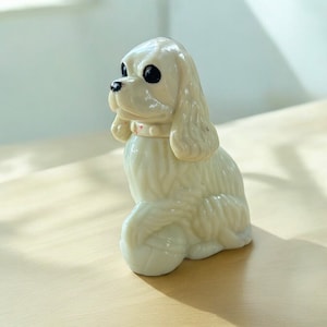 Puede incluir: Figura de cerámica blanca de un perro sentado con orejas caídas y collar. El perro está en posición sentada con la cabeza inclinada hacia un lado. La figura está hecha de cerámica blanca lisa y tiene un acabado brillante.