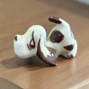 Könnte beinhalten: Eine Keramikfigur eines braunen und weißen Hundes in einer verspielten Pose. Der Hund liegt auf der Seite, der Kopf ist nach hinten geneigt und der Schwanz ist eingerollt.