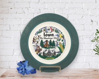 May include: A green and gold decorative plate with a white center featuring a colorful illustration of landmarks and symbols of Iowa, including the state capitol building, a church, a cornfield, and a cow. The text "Iowa The Hawkeye State" is printed on the plate.
