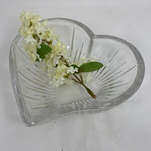 Op de afbeelding: Een heldere glazen kom in hartvorm met een geslepen glasontwerp. Een takje witte bloemen ligt in de kom.