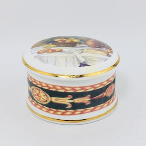May include: A white ceramic trinket box with a gold trim and a colorful image of fruit on the lid. The box has a decorative band around the side with a black, gold, and red design.