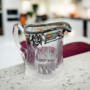 May include: A clear glass jug with a silver and orange design. The jug has a textured surface and a handle.